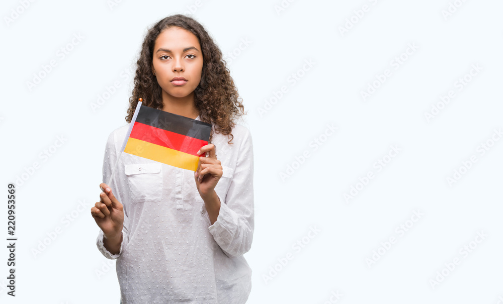 Young hispanic woman holding flag of Germany with a confident expression on smart face thinking serious Young hispanic woman holding flag of Germany with a confident expression on smart face thinking serious