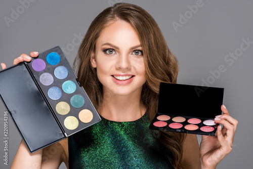 Photography portrait of smiling woman with eyeshadows palettes isolated on grey