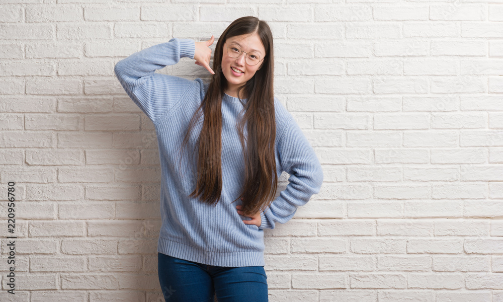 Young Chinise woman over white brick wall smiling doing phone gesture with hand and fingers like talking on the telephone. Communicating concepts. Young Chinise woman over white brick wall smiling doing phone gesture with hand and fingers like talking on the telephone. Communicating concepts.