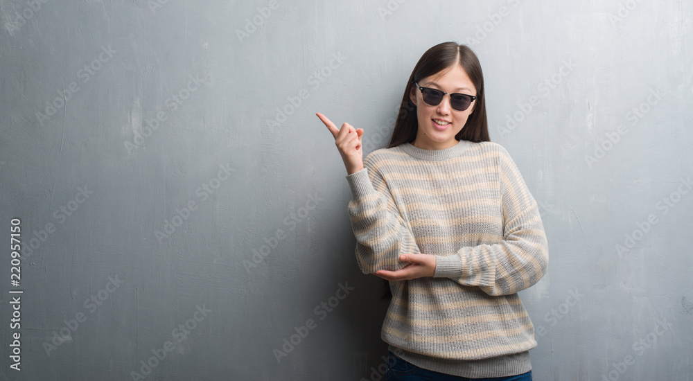 Young Chinese woman over grey wall wearing sunglasses very happy pointing with hand and finger to the side Young Chinese woman over grey wall wearing sunglasses very happy pointing with hand and finger to the side