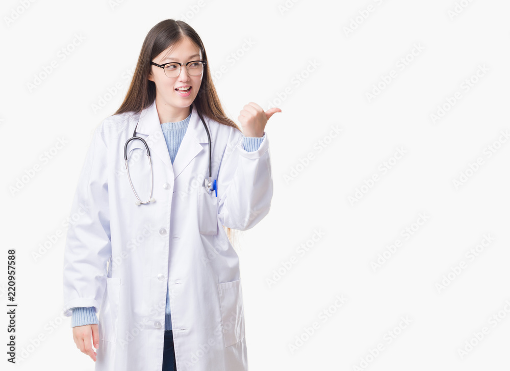 Young Chinese doctor woman over isolated background smiling with happy face looking and pointing to the side with thumb up. Young Chinese doctor woman over isolated background smiling with happy face looking and pointing to the side with thumb up.