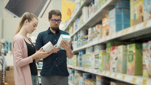 Married couple is buying baby formula in supermarket. They are standing near shelf and taking different jars from it, reading ingredients