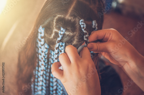 Hairdresser weaves braids with kanekalon material to young girl head, making creative hairstyle with thick plaits or pigtails also known as Afro braids photo