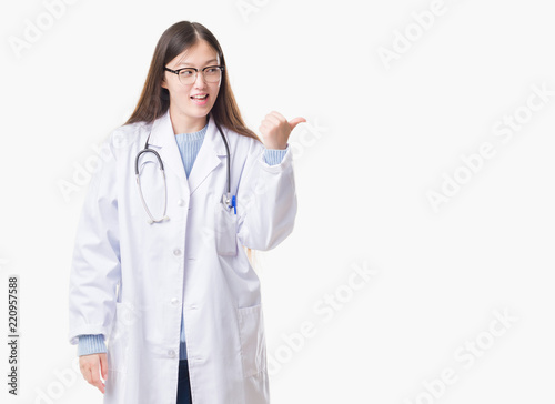 Young Chinese doctor woman over isolated background smiling with happy face looking and pointing to the side with thumb up.