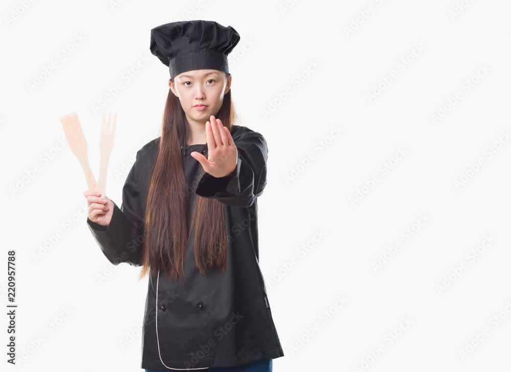 Young Chinese woman over isolated background wearing chef uniform with open hand doing stop sign with serious and confident expression, defense gesture Young Chinese woman over isolated background wearing chef uniform with open hand doing stop sign with serious and confident expression, defense gesture