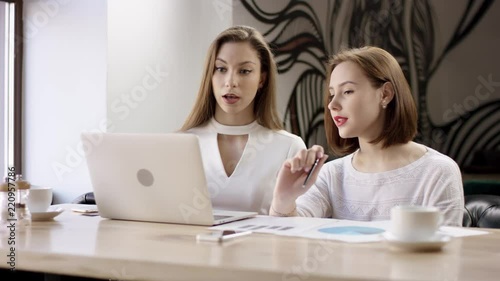 Two young business women are working with a laptop in a cafe. They are discussing and preparing the contract. It is a business meeting. Girls are sitting in the cafe at the table during coffee break
