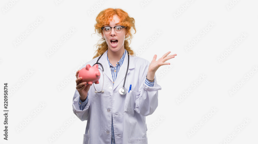 Young redhead doctor woman holding piggy bank very happy and excited, winner expression celebrating victory screaming with big smile and raised hands Young redhead doctor woman holding piggy bank very happy and excited, winner expression celebrating victory screaming with big smile and raised hands