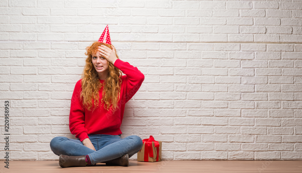 Young redhead woman sitting over brick wall wearing birthday hat stressed with hand on head, shocked with shame and surprise face, angry and frustrated. Fear and upset for mistake. Young redhead woman sitting over brick wall wearing birthday hat stressed with hand on head, shocked with shame and surprise face, angry and frustrated. Fear and upset for mistake.