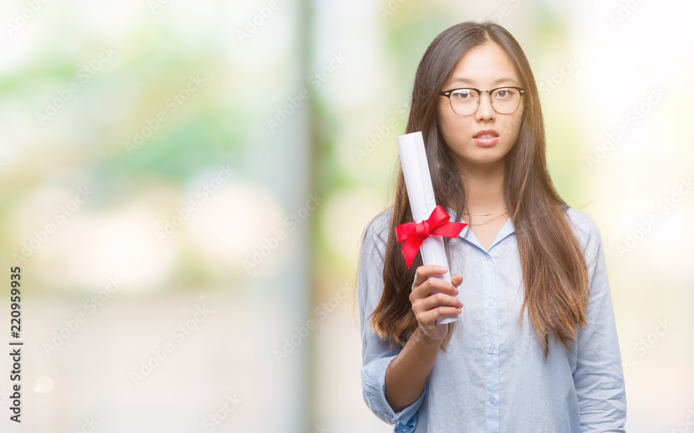 Young asian woman holding degree over isolated background with a confident expression on smart face thinking serious Young asian woman holding degree over isolated background with a confident expression on smart face thinking serious