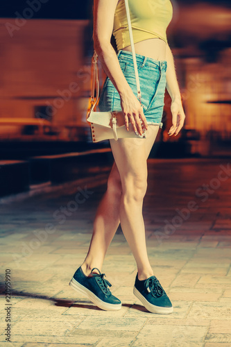 Night portrait of girl model wear on jeans shorts with small white handbag clutch, against lights of city streets.
