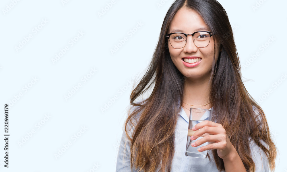 Young asian woman drinking a glass of water over isolated background with a happy face standing and smiling with a confident smile showing teeth Young asian woman drinking a glass of water over isolated background with a happy face standing and smiling with a confident smile showing teeth