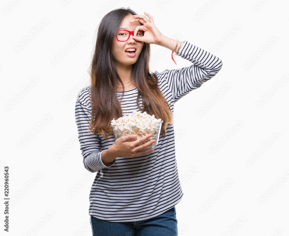 Young asian woman eating popcorn over isolated background with happy face smiling doing ok sign with hand on eye looking through fingers Young asian woman eating popcorn over isolated background with happy face smiling doing ok sign with hand on eye looking through fingers