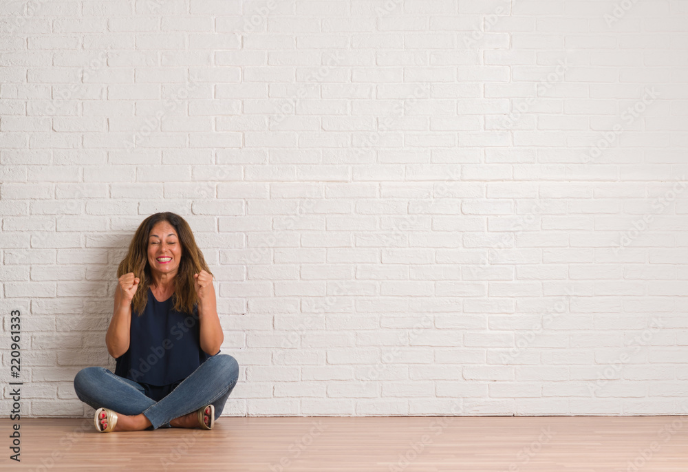 Middle age hispanic woman sitting on the floor over white brick wall excited for success with arms raised celebrating victory smiling. Winner concept. Middle age hispanic woman sitting on the floor over white brick wall excited for success with arms raised celebrating victory smiling. Winner concept.