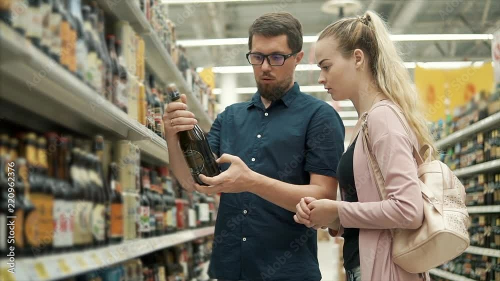 Pair is reading label and examining bottle with beer in a grocery. Man is rotating glass bottle and speaking with his girlfriend Pair is reading label and examining bottle with beer in a grocery. Man is rotating glass bottle and speaking with his girlfriend