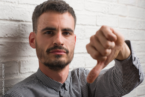 Young adult man standing over white brick wall with angry face, negative sign showing dislike with thumbs down, rejection concept