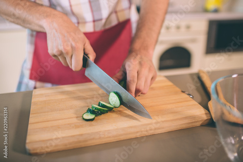 Close up of man's hands holding knife and cutting cucumber into pieces. He does it on desk. There is a glass bowl at table. Guy wears pron.