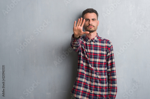 Young adult man standing over grey grunge wall with open hand doing stop sign with serious and confident expression, defense gesture
