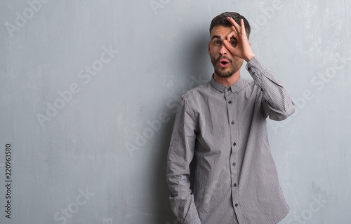 Young adult man standing over grey grunge wall doing ok gesture shocked with surprised face, eye looking through fingers. Unbelieving expression.