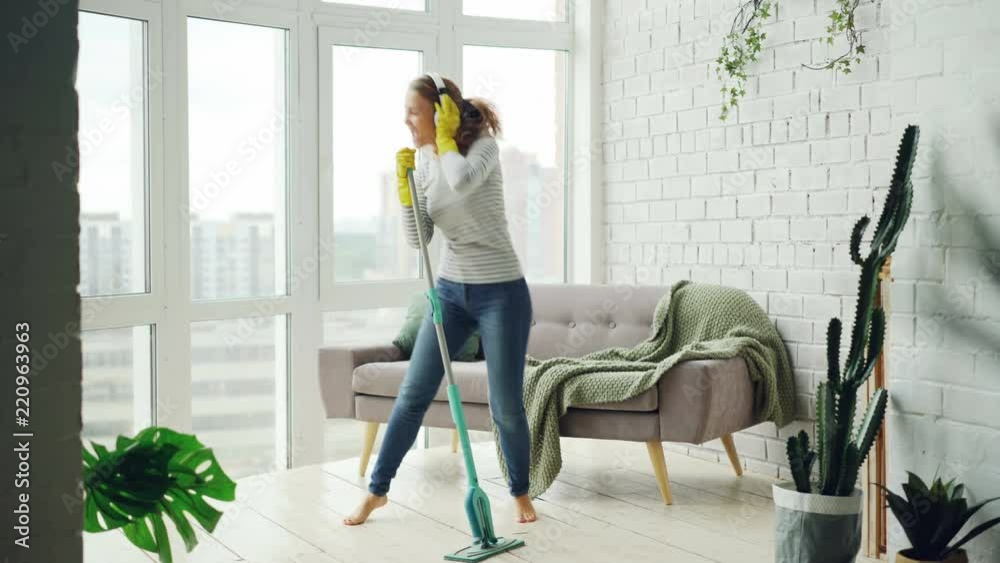Happy housewife is dancing and singing during housecleaning, she is listening to music in headphones and mopping floor having fun. Housework and technology concept.
