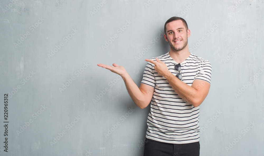 Young caucasian man over grey grunge wall amazed and smiling to the camera while presenting with hand and pointing with finger. Young caucasian man over grey grunge wall amazed and smiling to the camera while presenting with hand and pointing with finger.