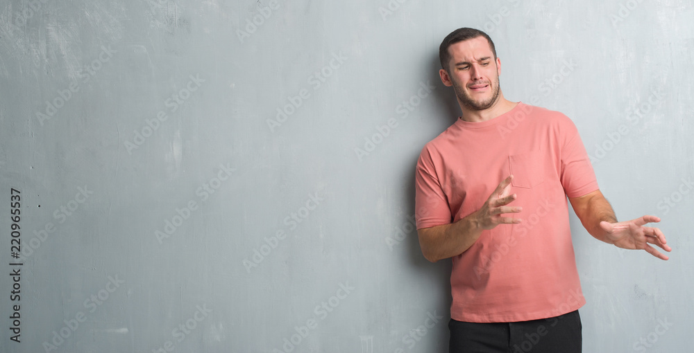 Young caucasian man over grey grunge wall disgusted expression, displeased and fearful doing disgust face because aversion reaction. With hands raised. Annoying concept.