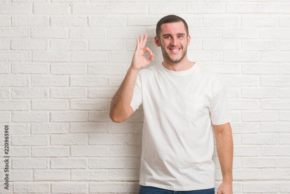 Young caucasian man standing over white brick wall smiling positive doing ok sign with hand and fingers. Successful expression. Young caucasian man standing over white brick wall smiling positive doing ok sign with hand and fingers. Successful expression.