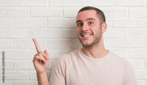 Young caucasian man standing over white brick wall very happy pointing with hand and finger to the side