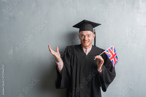 Young redhead man over grey grunge wall wearing graduate uniform holding united kigdom flag very happy and excited, winner expression celebrating victory screaming with big smile and raised hands photo