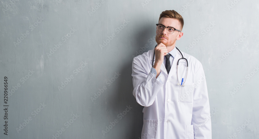 Young redhead doctor man over grey grunge wall serious face thinking about question, very confused idea Young redhead doctor man over grey grunge wall serious face thinking about question, very confused idea