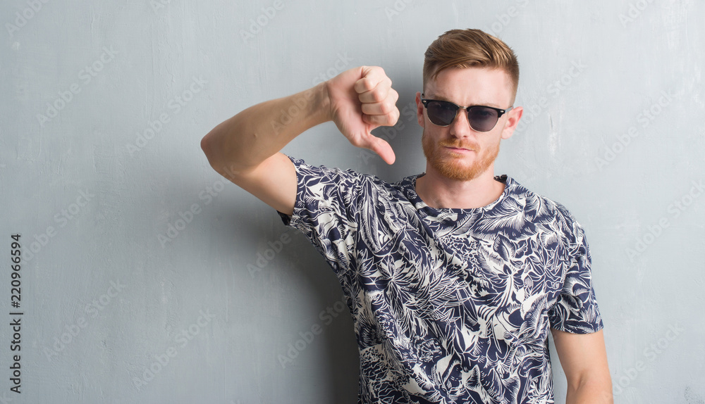 Young redhead man over grey grunge wall wearing sunglasses with angry face, negative sign showing dislike with thumbs down, rejection concept Young redhead man over grey grunge wall wearing sunglasses with angry face, negative sign showing dislike with thumbs down, rejection concept