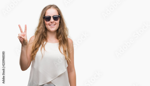 Young blonde woman wearing sunglasses showing and pointing up with fingers number two while smiling confident and happy.