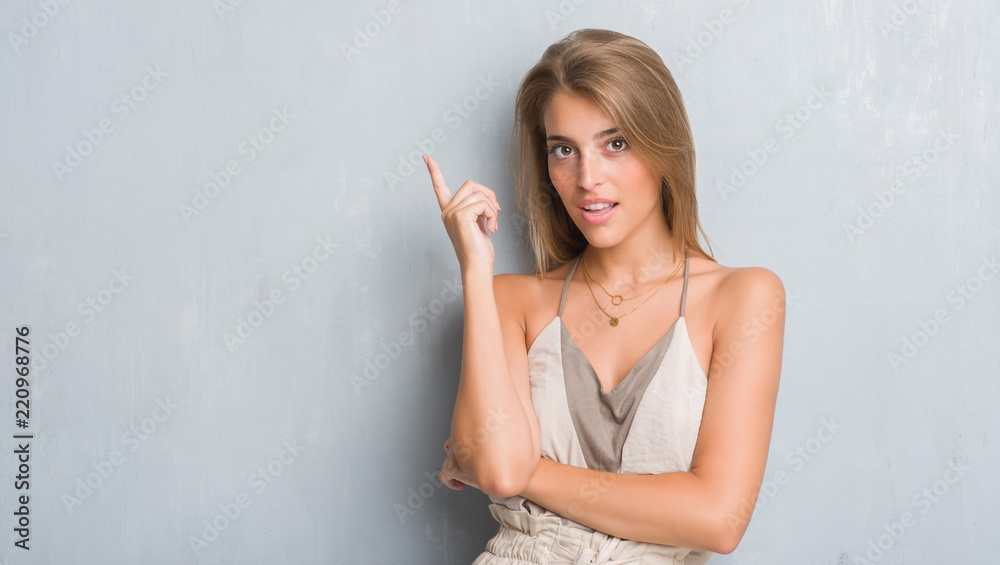 Beautiful young woman standing over grunge grey wall surprised with an idea or question pointing finger with happy face, number one Beautiful young woman standing over grunge grey wall surprised with an idea or question pointing finger with happy face, number one