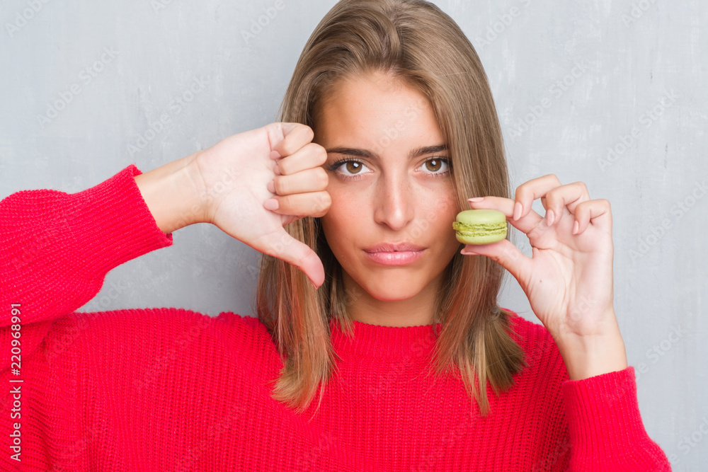 Beautiful young woman over grunge grey wall eating green macaron with angry face, negative sign showing dislike with thumbs down, rejection concept Beautiful young woman over grunge grey wall eating green macaron with angry face, negative sign showing dislike with thumbs down, rejection concept