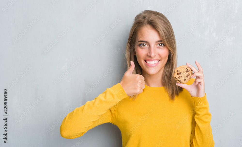 Beautiful young woman over grunge grey wall eating chocolate chip cooky happy with big smile doing ok sign, thumb up with fingers, excellent sign Beautiful young woman over grunge grey wall eating chocolate chip cooky happy with big smile doing ok sign, thumb up with fingers, excellent sign