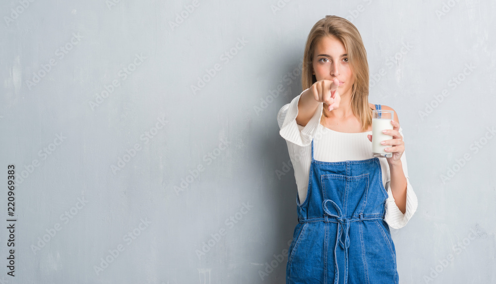 Beautiful young woman over grunge grey wall driking a glass of milk pointing with finger to the camera and to you, hand sign, positive and confident gesture from the front Beautiful young woman over grunge grey wall driking a glass of milk pointing with finger to the camera and to you, hand sign, positive and confident gesture from the front