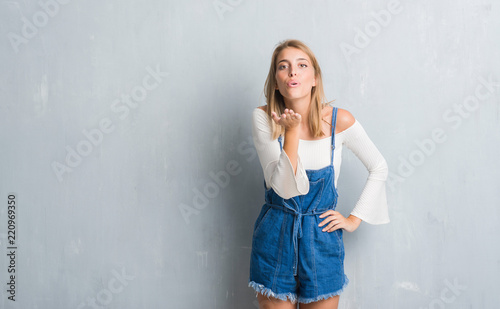 Beautiful young woman standing over grunge grey wall looking at the camera blowing a kiss with hand on air being lovely and sexy. Love expression.