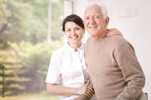 Friendly smiling caregiver hugging happy elderly man in the hospital