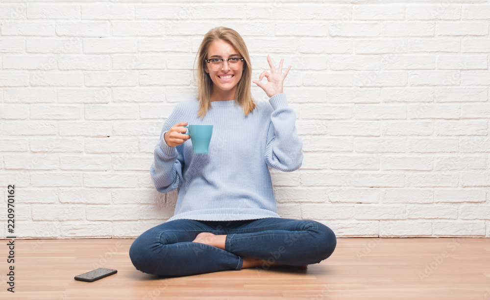 Beautiful young woman sitting on the floor driking cup of coffee at home doing ok sign with fingers, excellent symbol Beautiful young woman sitting on the floor driking cup of coffee at home doing ok sign with fingers, excellent symbol