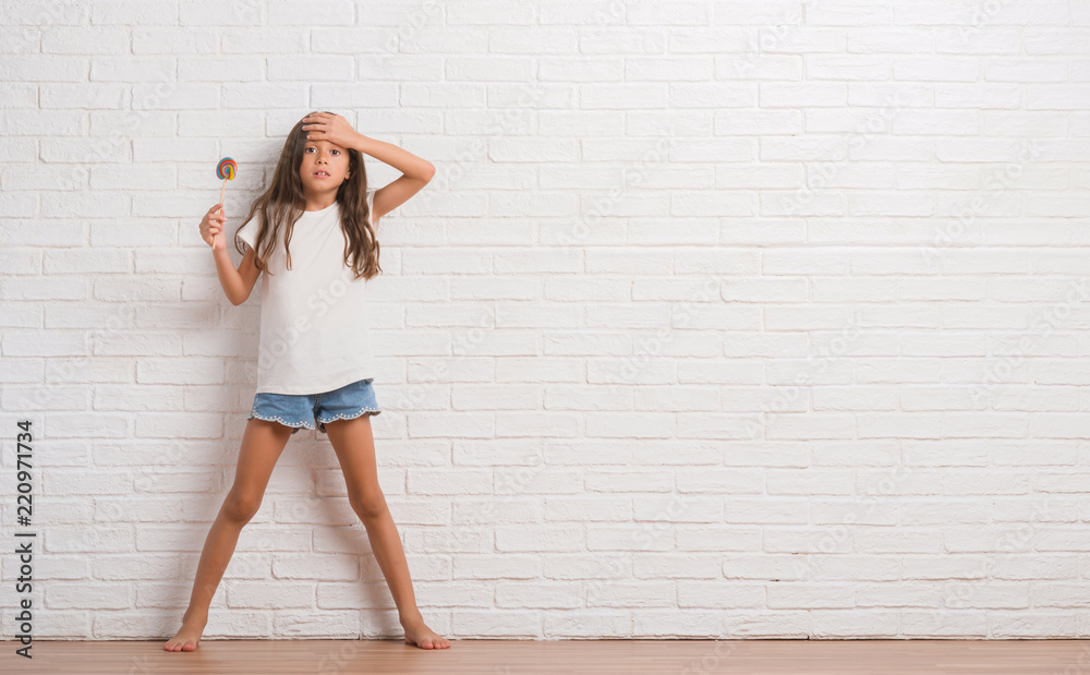 Young hispanic kid over white brick wall eating lollipop candy at home stressed with hand on head, shocked with shame and surprise face, angry and frustrated. Fear and upset for mistake.