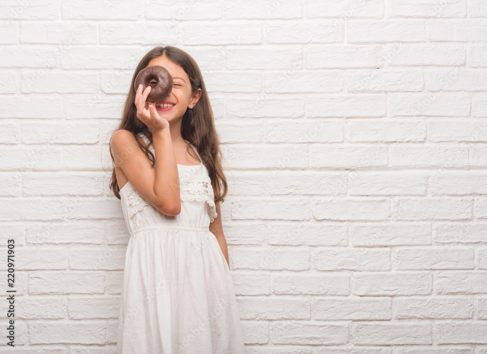 Young hispanic kid over white brick wall eating chocolate donut with a happy face standing and smiling with a confident smile showing teeth Young hispanic kid over white brick wall eating chocolate donut with a happy face standing and smiling with a confident smile showing teeth