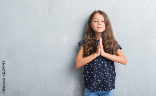 Young hispanic kid over grunge grey wall praying with hands together asking for forgiveness smiling confident.