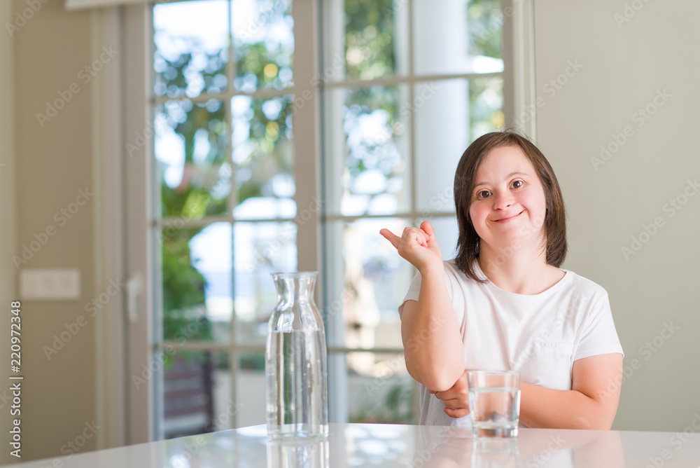 Down syndrome woman at home drinking water very happy pointing with hand and finger to the side Down syndrome woman at home drinking water very happy pointing with hand and finger to the side
