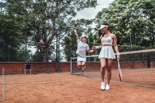 Little champion on tennis court! Full length image of jumping child tennis player with sporty trainer. Tennis coach holds hand of little girl celebrating victory. Active leisure outdoors.