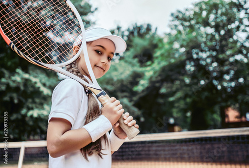 Tennis training for young kid outdoors. Portrait of happy sporty little girl on tennis court. Caucasian child in white tennis sportswear on training. © HBS