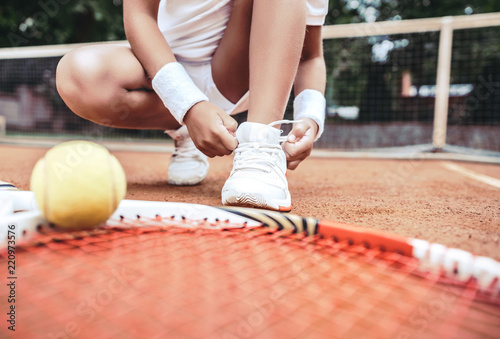 Girl on tennis court in the sport club. Cropped image of a little tennis player. Girl child tying shoelaces on tennis court. Summer activities for children. © HBS