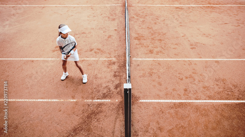 Child playing tennis on outdoor clay court. Top view of little tennis player on open tennis court. Full length shot of sporty child girl on tennis training in the club. Active exercise for kids. © HBS