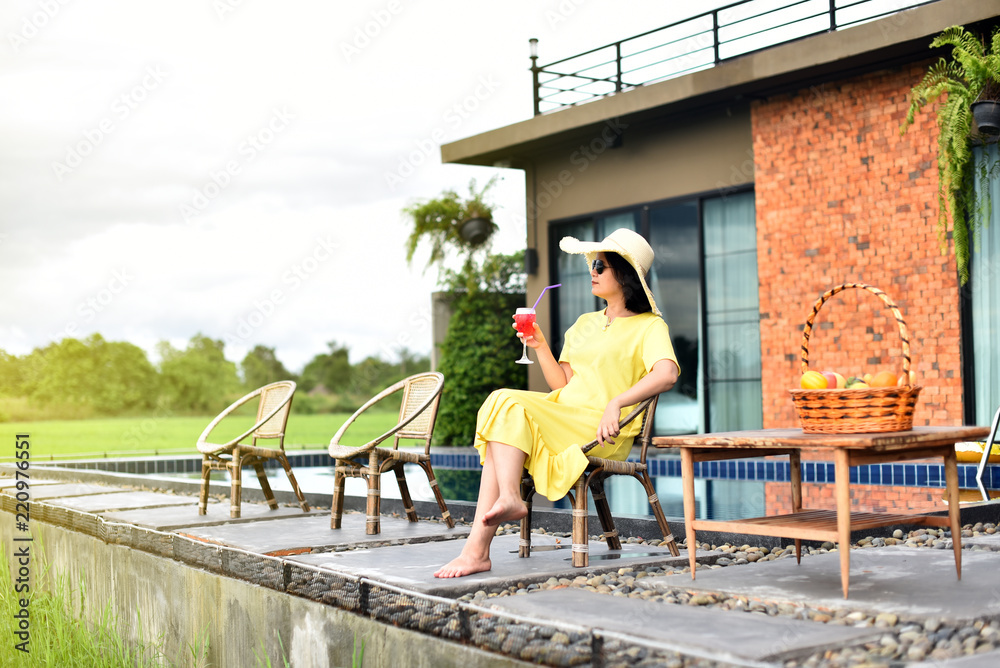 Portrait of Asian woman wear yellow dress sitting near swimming pool and holding cocktail in hand
