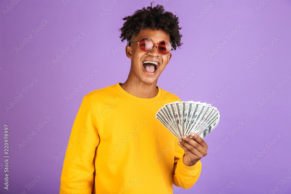 Portrait of fashion african american man wearing sunglasses smiling while holding cash money dollars, isolated over violet background Portrait of fashion african american man wearing sunglasses smiling while holding cash money dollars, isolated over violet background