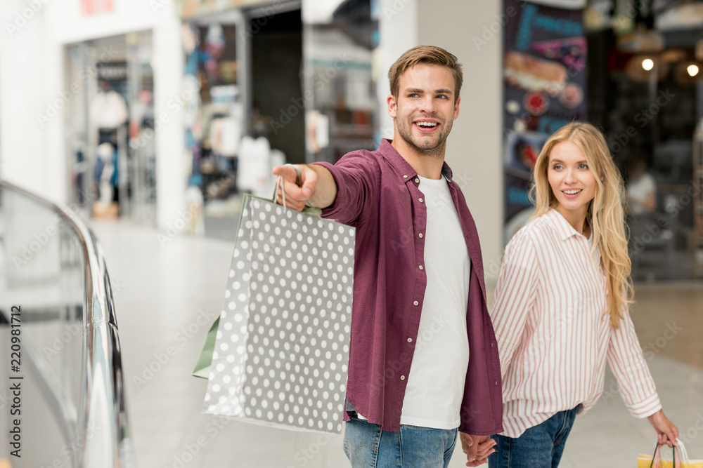 selective focus of smiling young man with shopping bags pointing to  girlfriend walking near at shopping mall Stock Photo | Adobe Stock