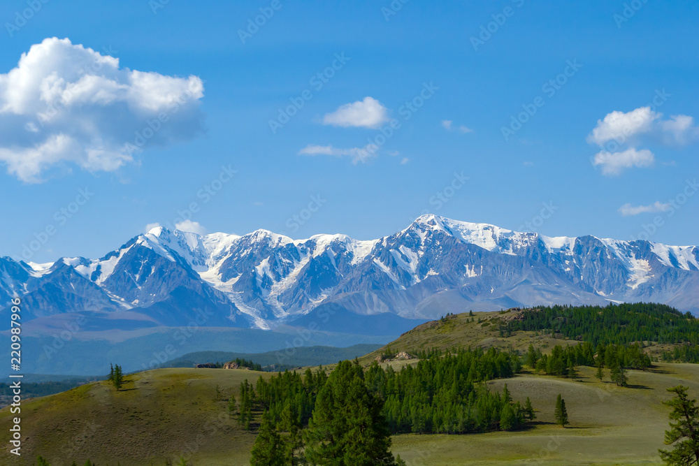 Landscape of snow-capped mountains, with green hilly valley a fresh summer day under a blue sky with white clouds and sun rays in Altai mountains